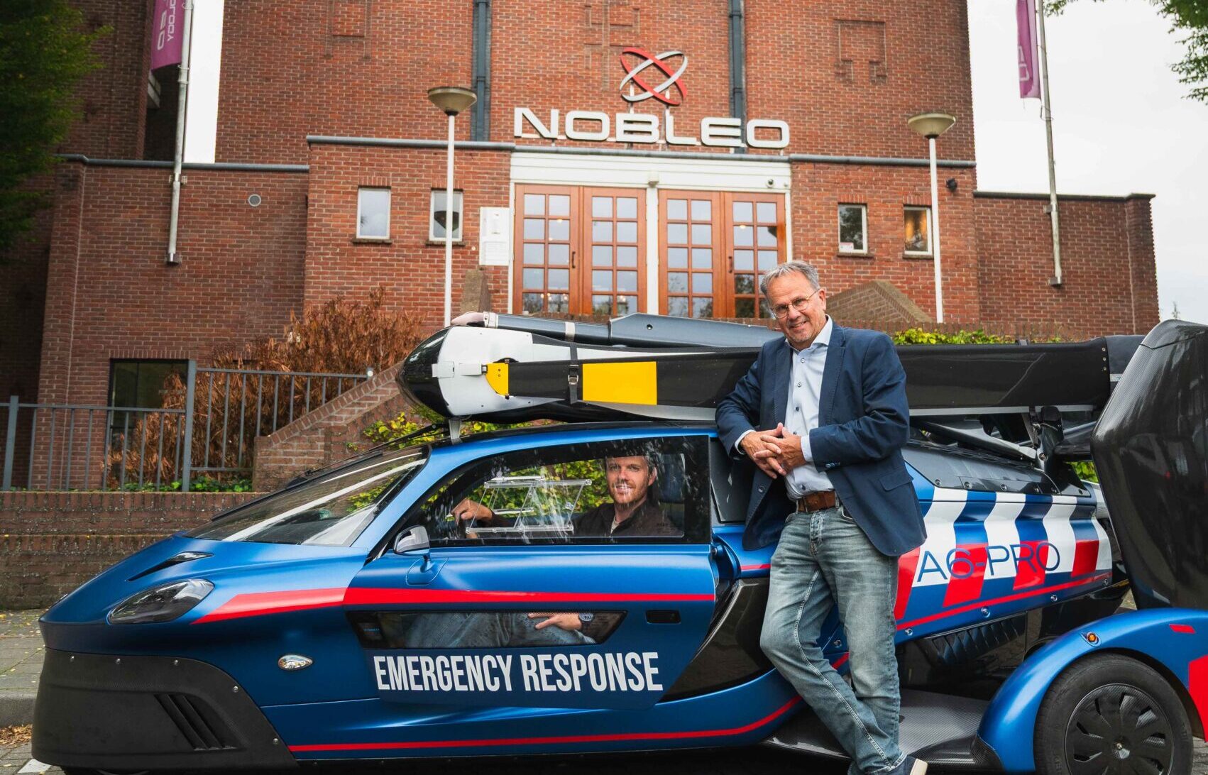 A person leaning against a PAL-V Liberty FlyDrive vehicle and a person in a PAL-V Liberty FlyDrive vehicle in front of the Nobleo Manufacturing building in Eindhoven